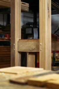 Close-up of a timber bus shelter component during assembly, showing traditional mortise and tenon joinery and wooden dowel pegs in a woodworking workshop.