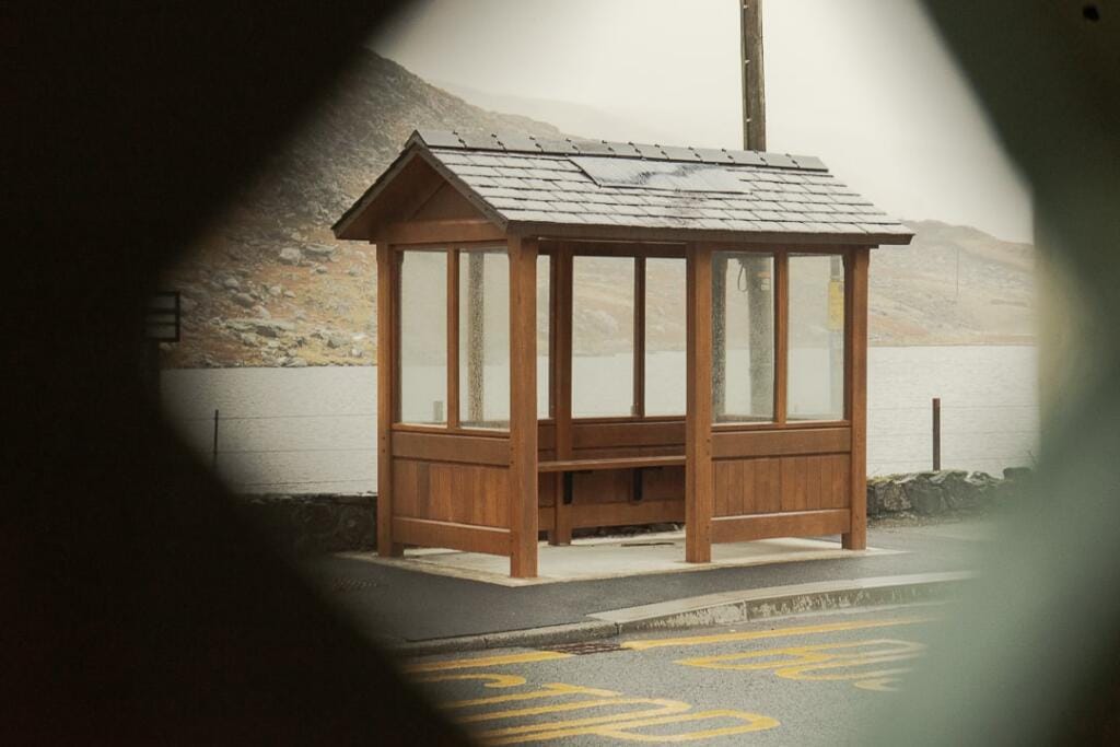 A handcrafted timber bus shelter set against a rural landscape with trees and grass, demonstrating durability and design in a real-world setting.