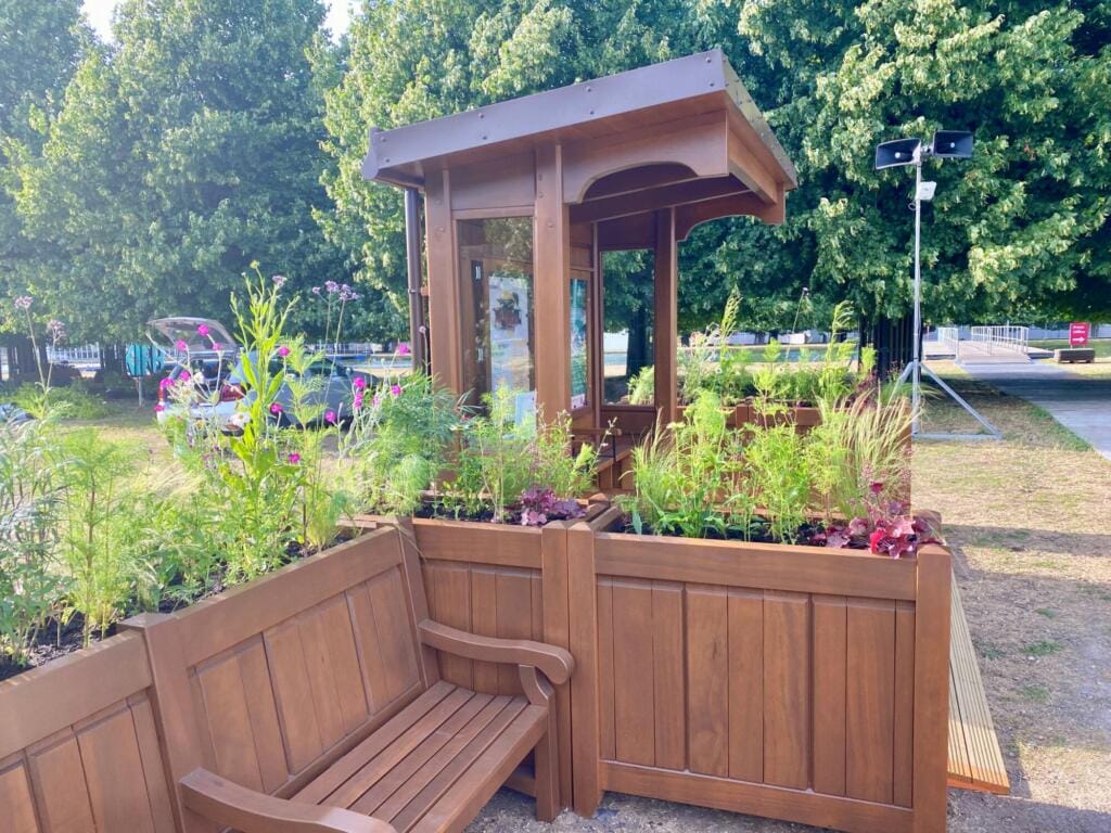 A handcrafted wooden shelter surrounded by tall planters and integrated seating, set against a backdrop of leafy trees at RHS Hampton Court Palace Garden Festival.